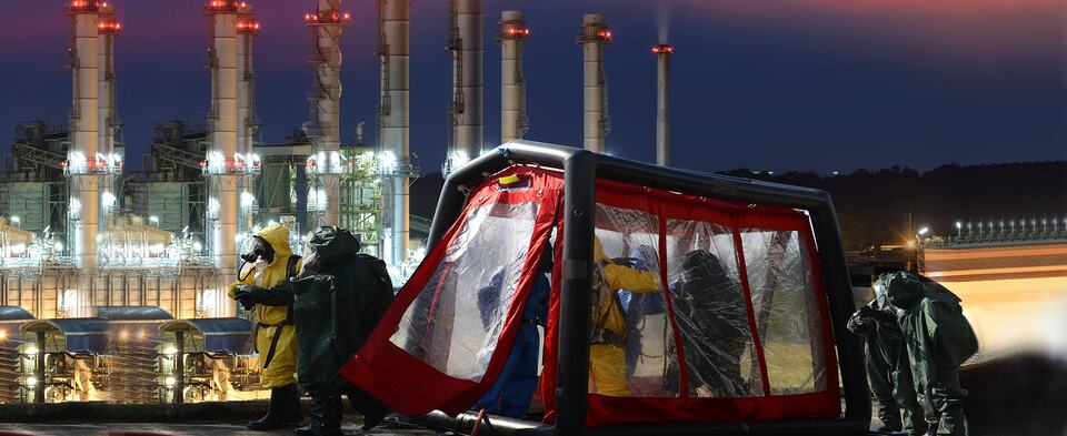Responders in protective suits beside a decontamination tent at an industrial site at dusk